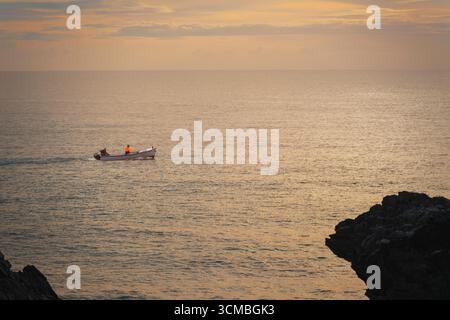 Early morning off Wicklow coast with two fishermen in a small dinghy, calm sea, and rocky headland in view. Stock Photo