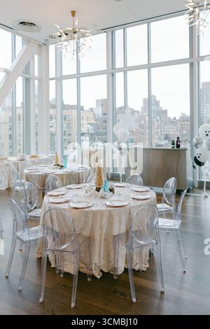 A bright event space features round tables with white tablecloths and transparent chairs, providing a city skyline view. Stock Photo