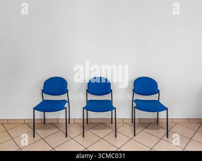 Three blue chairs in a minimalist style against a white wall and brown tile floor in the waiting room. Copy space Stock Photo