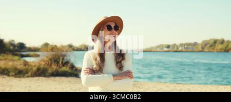 Stylish mature woman on coast beach, fashionable middle aged female model posing outdoors, wearing sweater, hat and glasses, standing with crossed arm Stock Photo