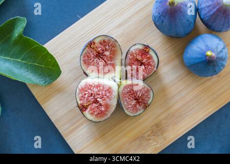 Fresh purple figs whole and sliced showing pink flesh on wooden cutting board with fig leaf. Healthy organic Mediterranean fruit flat. Stock Photo