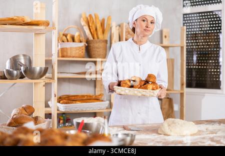 Portrait of happy female baker in white uniform working in bakehouse, holding tray with fresh bakery goods on kitchen Stock Photo