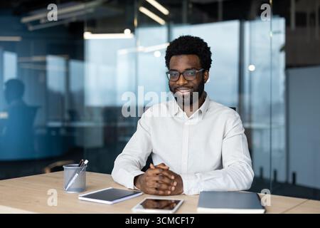 Confident african american entrepreneur smiling while seated at a desk in a bright, modern office, radiating professionalism and success in a dynamic Stock Photo
