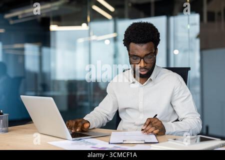 Young african american entrepreneur using laptop and taking notes while sitting at desk and working with documents and charts in contemporary office Stock Photo