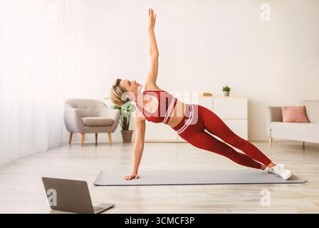 Middle-aged woman doing side plank on mat with pc Stock Photo
