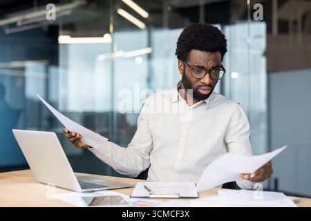 Young businessman looking puzzled while reviewing documents and working on a laptop at his desk in a sleek, modern office environment, surrounded by g Stock Photo