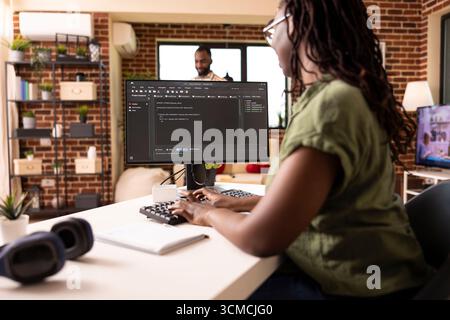 Remote female programmer works from cozy home desk, coding for startup project on desktop pc. Tech savvy black woman managing web development tasks on computer in brick wall apartment. Stock Photo