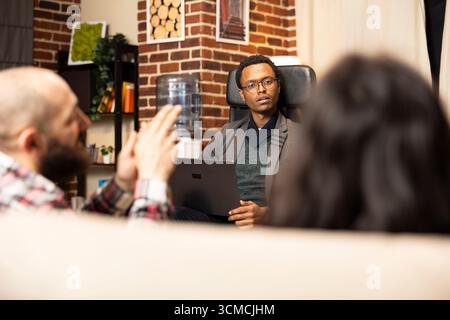 African American psychotherapist sits across from young couple, listening and documenting counseling session on laptop. Black male counselor offers professional guidance to married clients. Stock Photo