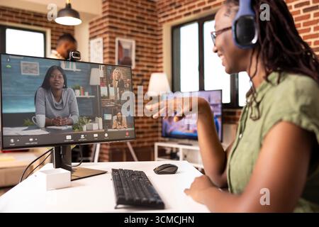 Professional female individual attending video conference using desktop pc and webcam. Remote worker manages virtual team meetings and company discussions from cozy brick wall home. Stock Photo