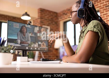 Young black woman sitting at desk in home office, engaging in online discussions with coworkers on desktop pc. Female freelancer talking virtually with group of shareholders on video call. Stock Photo