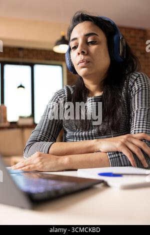 Female freelancer with wireless headset watching online business lecture on laptop. Serious South Asian woman attends virtual webinar, listening attentively and learning remotely from home. Stock Photo
