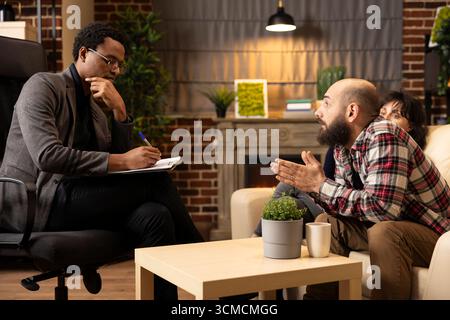 Diverse individuals participating in ongoing therapy session. Black counselor listening attentively and taking notes on clipboard as white male patient discusses relationship issues. Stock Photo