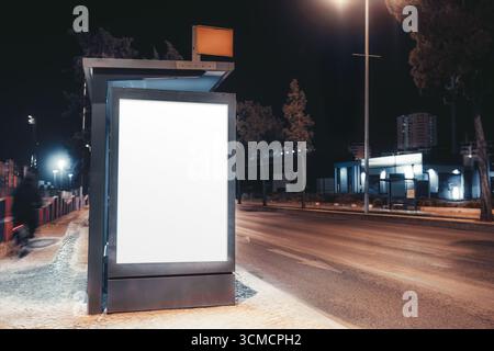 Blank illuminated billboard at bus stop on empty city street at night, glowing under streetlights, ready for advertising, urban marketing, promotion, Stock Photo
