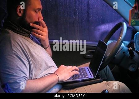 Hispanic male traveler using a laptop sitting in his car, it is nighttime and raining outside. Concepts: digital nomad, remote work and working while Stock Photo