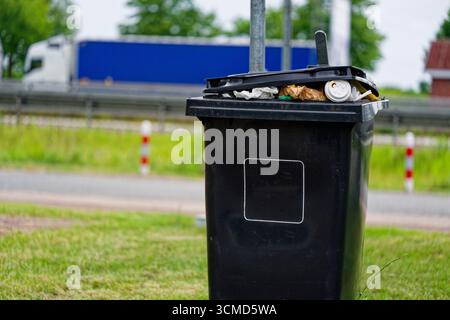 A black trash bin is overfull and the lid sits ajar. Paper bags from a bakery poke out beside disposable coffee cups and mixed litter. Behind it stret Stock Photo