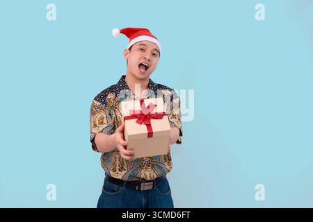 young happy man wearing batik shirt and santa hat giving gift box in christmas concept blue studio background Stock Photo