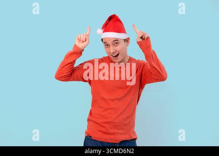 A cheerful young man wearing a red sweater and Santa hat points upward in christmas concept blue background Stock Photo