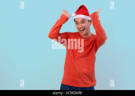cheerful young man with santa hat poses playfully in red shirt against blue studio background Stock Photo