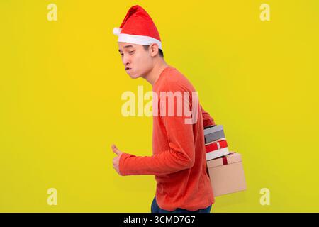 Cheerful man in a Santa hat and red shirt carries several gift boxes on his back, smiling against a vibrant yellow background Stock Photo