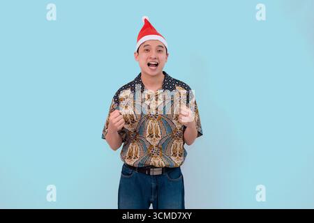 A cheerful young man wearing traditional batik with Santa hat against a light blue background expressing excitement and holiday spirit Stock Photo
