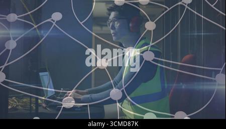 Technician wearing vest, headphones using laptop at data center, with server racks, network cables Stock Photo