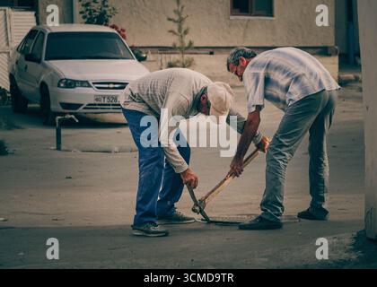 Two adult Uzbek men work hard breaking pavement with a pickaxe in Bukhara, Uzbekistan Stock Photo