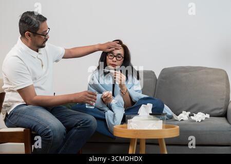 A caring man taking care of a sick woman wrapped in blanket, checking her fever with thermometer while she sits on sofa, showing healthcare, illness, Stock Photo