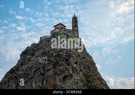 Low angle view of Saint Michel’s church perched atop a volcanic rock in Le Puy-en-Velay, set against a vivid blue sky with puffy white clouds. Stock Photo