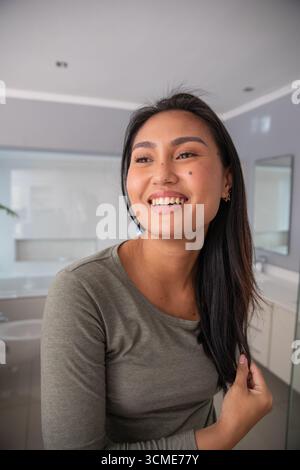 Mid adult Asian woman standing in modern home bathroom touching long dark hair beside white vanity Stock Photo