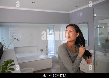 Asian woman brushing long dark hair in modern bathroom by vanity with potted plants, copy space Stock Photo