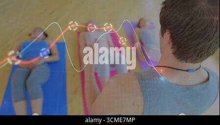Leading instructor guiding group lying on mats on studio floor, wearing sensor showing data overlay Stock Photo