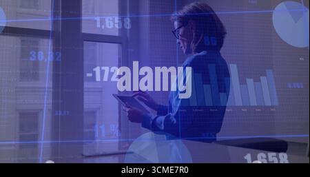 Professional woman in blue blouse analyzing financial charts near office window, with tablet graphs Stock Photo