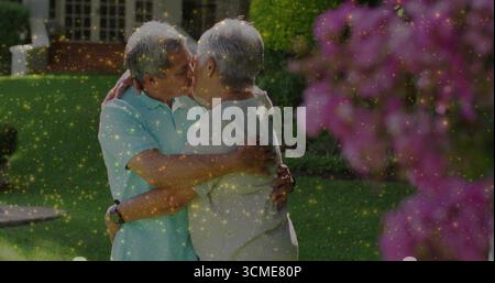 Embracing senior couple sharing gentle kiss in front yard, with flowering pink shrub and green lawn Stock Photo