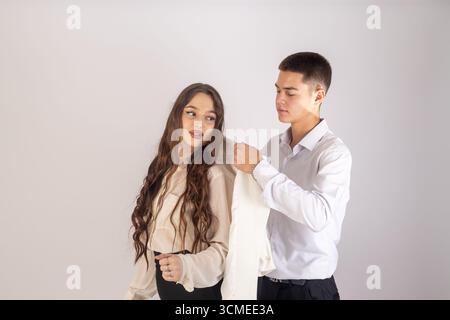 man is helping a young woman with long, dark hair put on a light-colored jacke Stock Photo