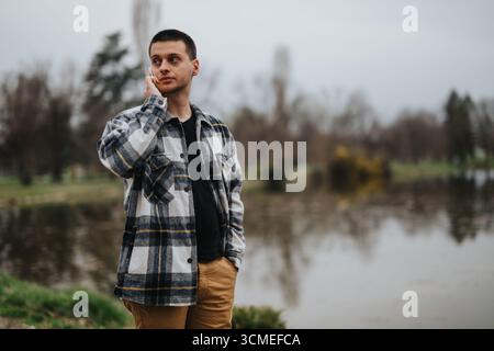 Man in plaid jacket talking on phone by a scenic wooded pond Stock Photo