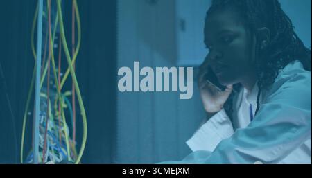 Tech wearing lab coat inspecting server rack, holding phone in data center with network cables Stock Photo