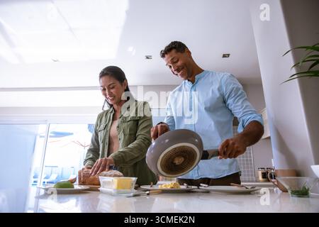 Diverse couple slicing bread and pouring eggs from frying pan at kitchen island with avocado Stock Photo