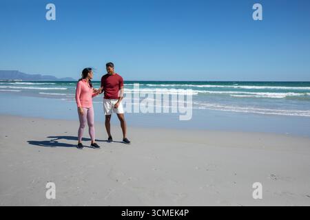 Diverse couple in athletic wear walking and talking on sandy beach next to waves, copy space Stock Photo