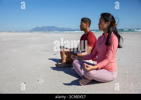 Diverse couple meditating in yoga pose on sandy beach at oceanfront in sportswear, copy space Stock Photo