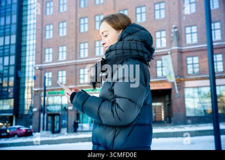 Middle aged woman walking on snowy city street with smartphone, urban winter lifestyle, modern technology, and authentic everyday life. Stock Photo