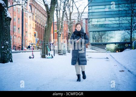Middle aged woman walking on snowy city street, checking smart watch, urban winter lifestyle, modern technology, professional and active day. Stock Photo