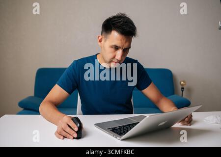 Portrait of freelancer male concentrating on work while sitting at white table in modern home office, using vertical mouse and laptop to enhance Stock Photo