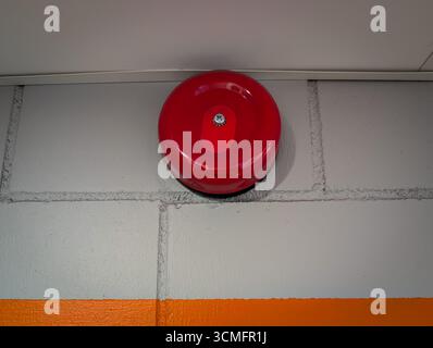 A classic red metal fire alarm bell for emergency alerts, mounted on a grey painted cinder block industrial wall. Stock Photo