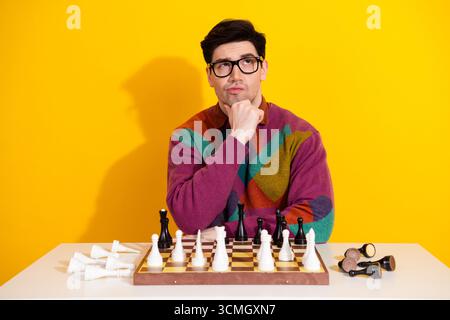 Thoughtful young man plays chess, contemplating his next move, wearing stylish attire against a yellow background Stock Photo