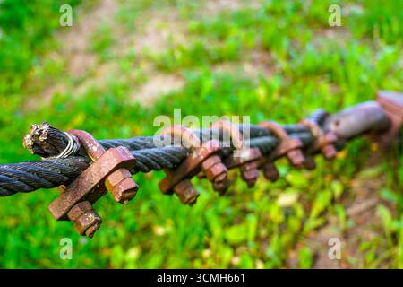 Thick wire rope fastening system with steel clamps and bolts on green natural background, industrial wire rope tension system outdoors Stock Photo
