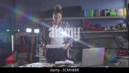 Examining printed plans on workshop workbench, mechanic using laptop with red car and notebook Stock Photo