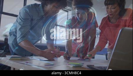 Collaborating team writing sticky notes on table near laptop at modern office, with digital overlay Stock Photo