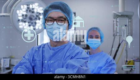 Standing surgeons wearing gowns and masks in operating room, showing lamp, monitors, icons overlay Stock Photo