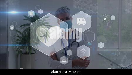 Standing senior doctor wearing lab coat, mask and stethoscope in hospital corridor, medical icons Stock Photo