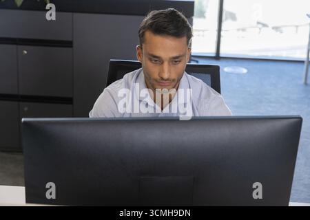 Hispanic male working at white desk in office using large monitor with mesh chair Stock Photo